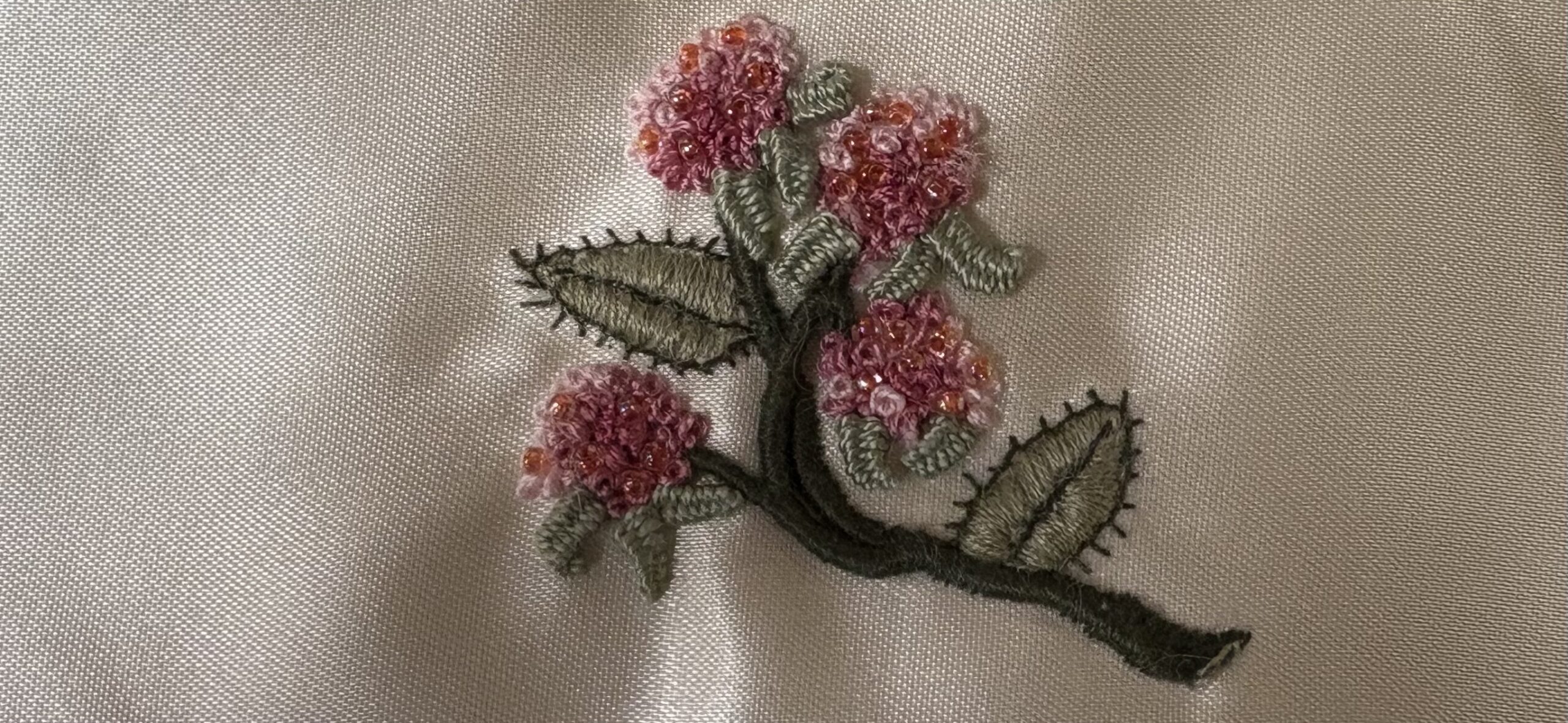 Embroidered branch with green leaves and clusters of pink flowers on a light fabric background.