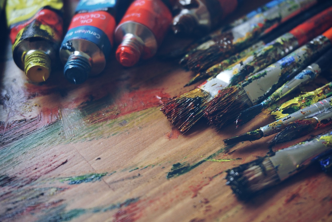 Tubes of paint and used paintbrushes lie on a messy, paint-splattered wooden surface.
