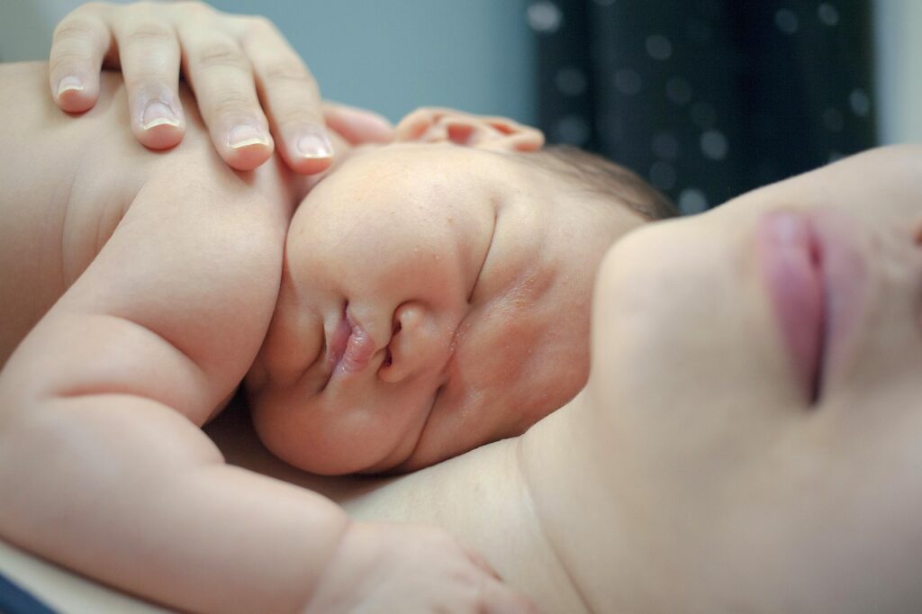 A newborn baby sleeps peacefully on an adult’s chest, gently held by a hand.