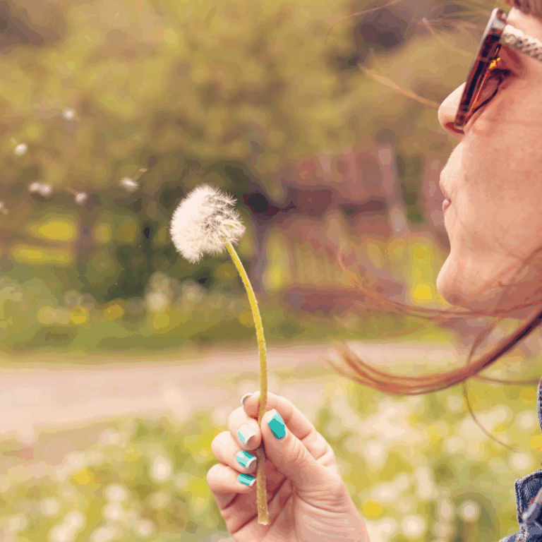 A person wearing sunglasses blows on a dandelion, scattering its seeds outdoors on a sunny day.