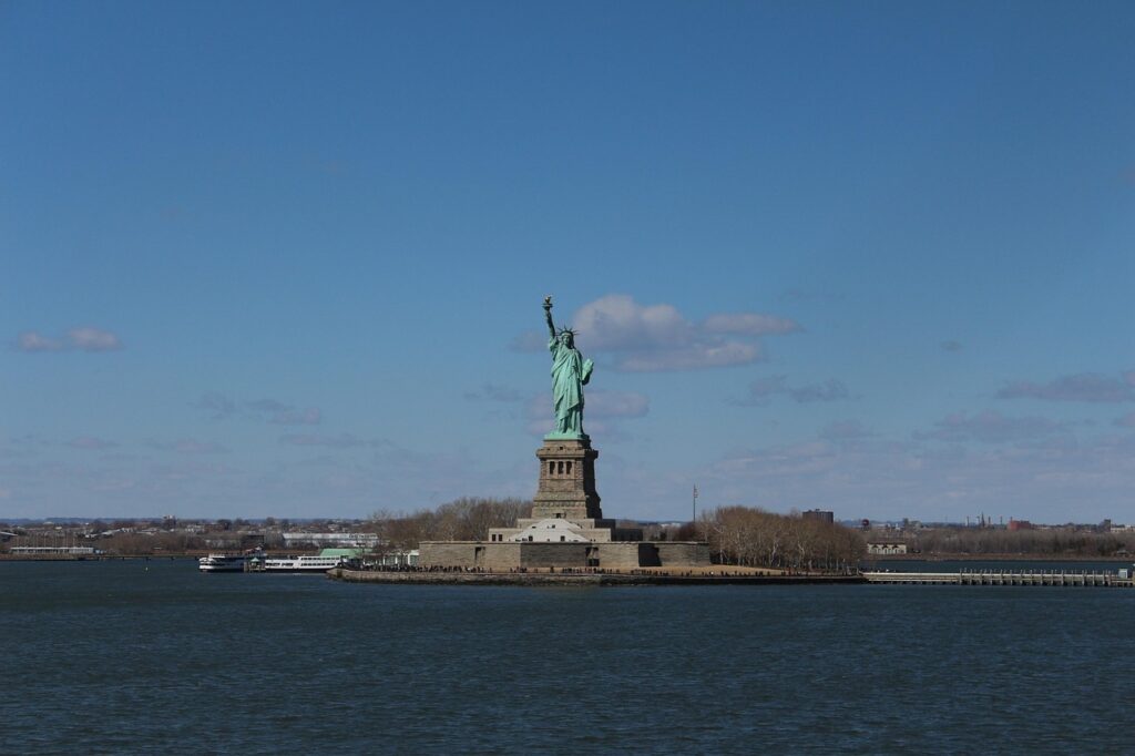 The Statue of Liberty stands on Liberty Island with a blue sky and water in the foreground.