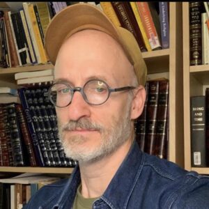 A man with glasses and a cap poses in front of bookshelves filled with books.