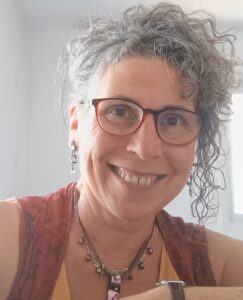 Smiling person with curly gray hair, glasses, and jewelry, sitting indoors in natural light.