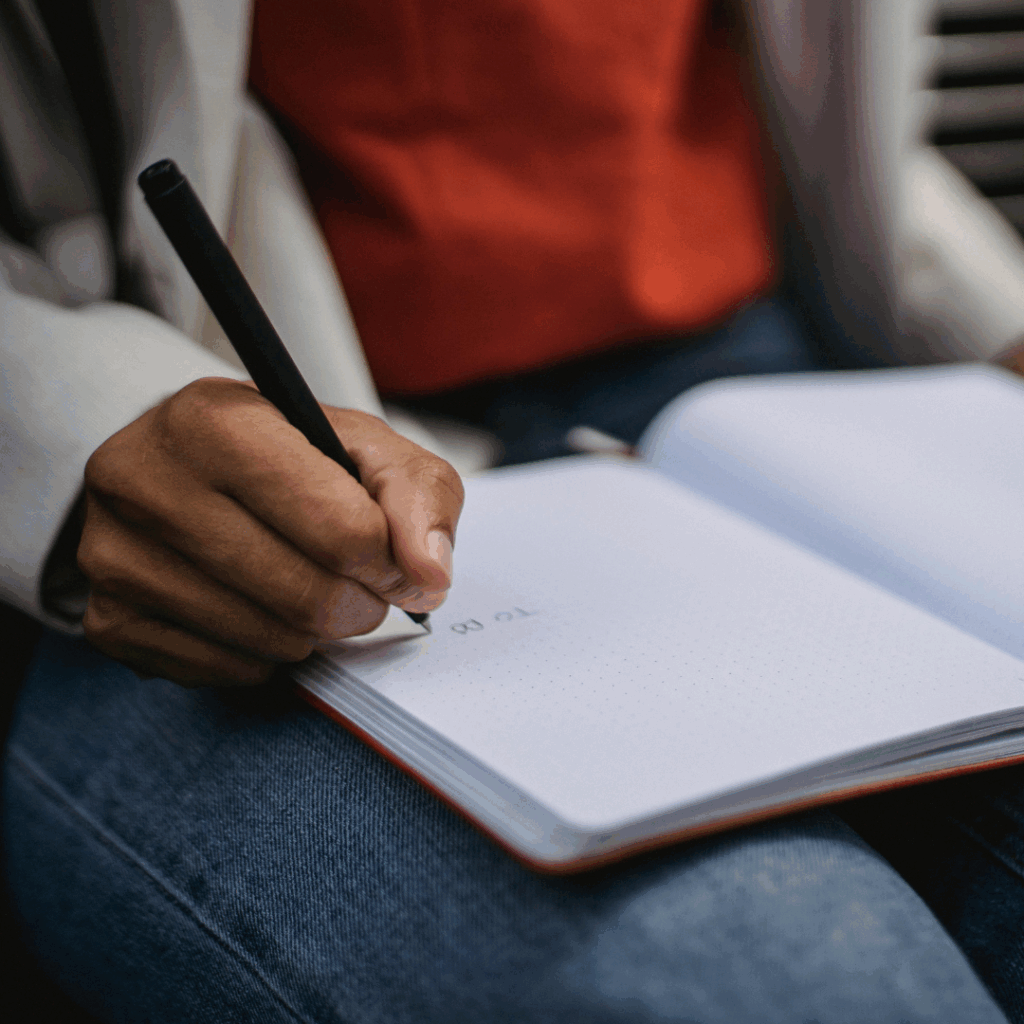 A person writes To do in a notebook with a pen, sitting and wearing jeans and a red top.