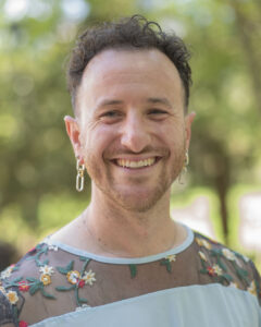 A person with short curly hair, earrings, and a sheer floral top smiles outdoors with greenery behind them.
