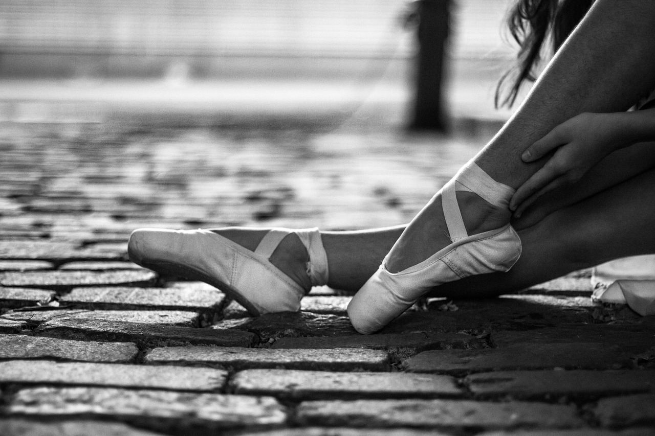Ballet dancer sitting on cobblestones, adjusting pointe shoes, shown in black and white.