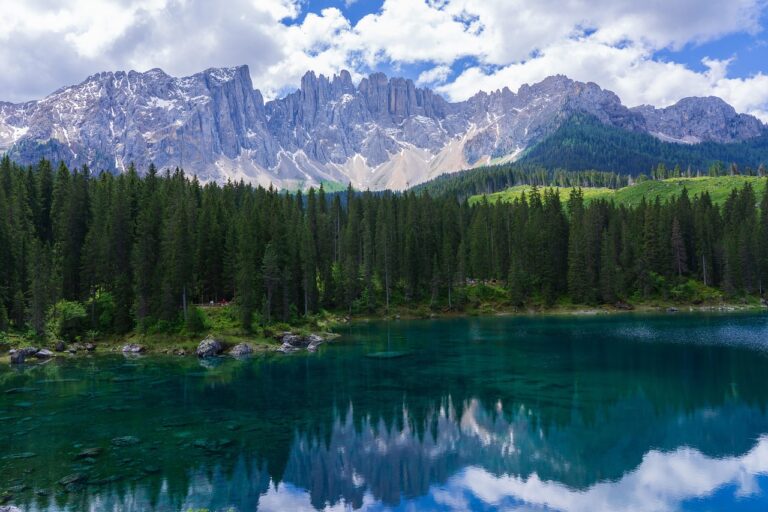 Clear blue lake reflects pine trees and rugged mountains under a partly cloudy sky.