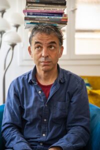 A man sits indoors with several books balanced on his head, looking upward thoughtfully.