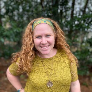 Smiling woman with curly red hair wearing a yellow lace top and headband, standing outdoors with trees behind her.