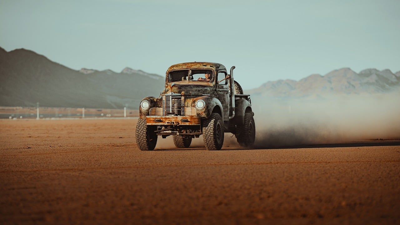 An old rusty pickup truck drives fast across a dusty desert with mountains in the background.