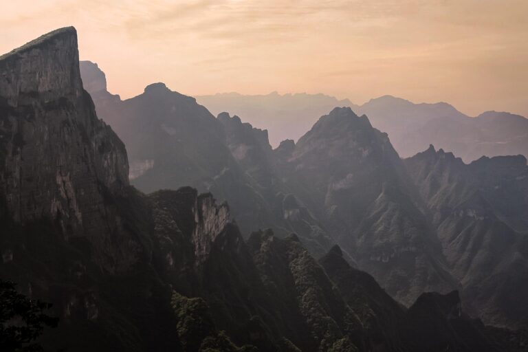 Jagged mountain peaks covered in greenery under a hazy, pastel-colored sky at sunset or sunrise.