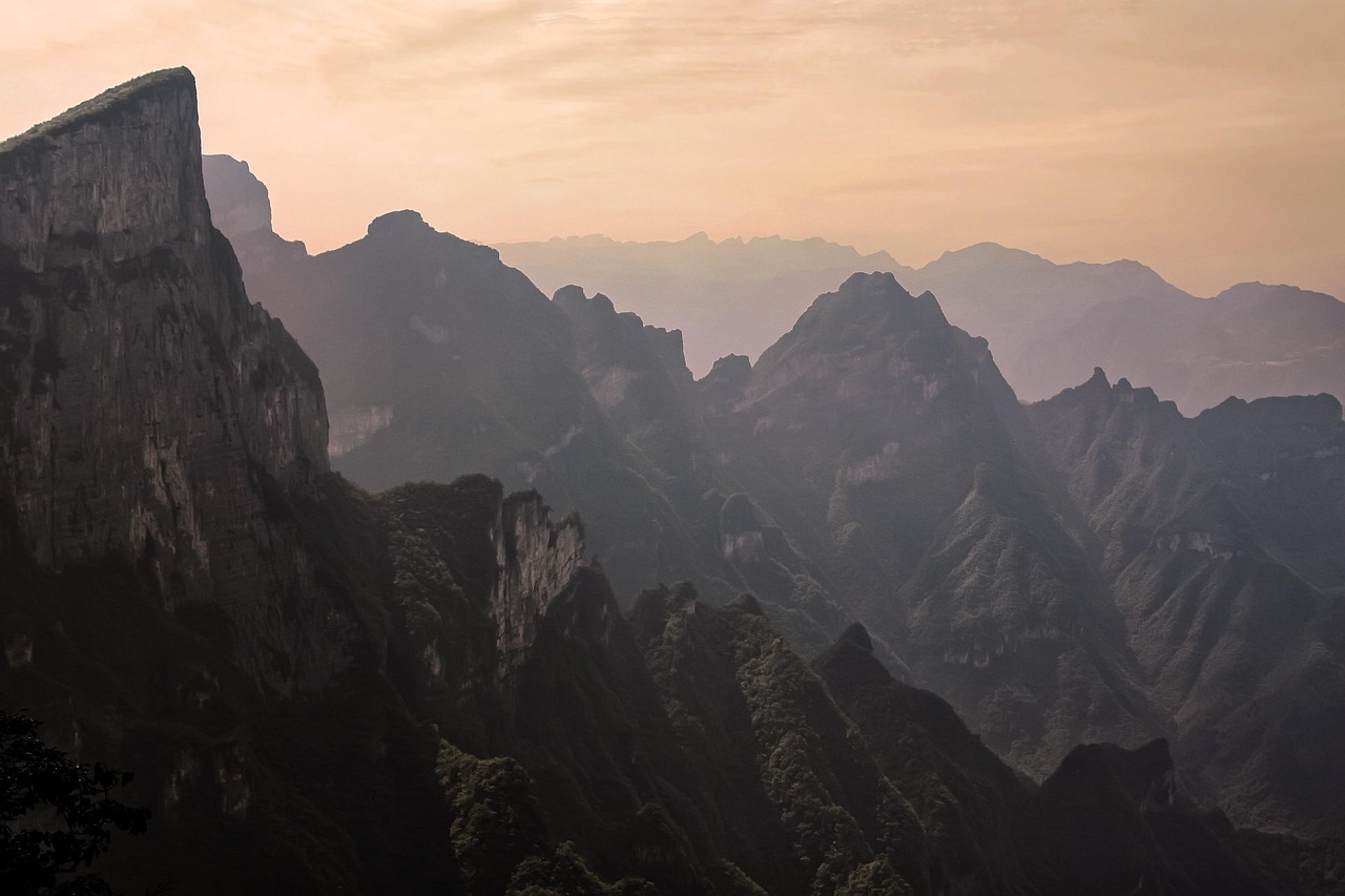 Jagged mountain peaks covered in greenery under a hazy, pastel-colored sky at sunset or sunrise.