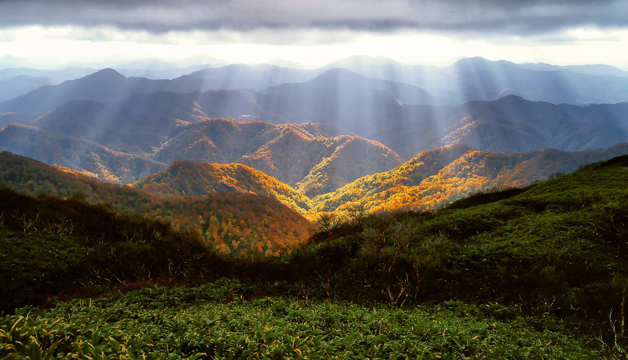 Sunbeams shine through clouds onto green and autumn-colored hills and mountains in a scenic landscape.