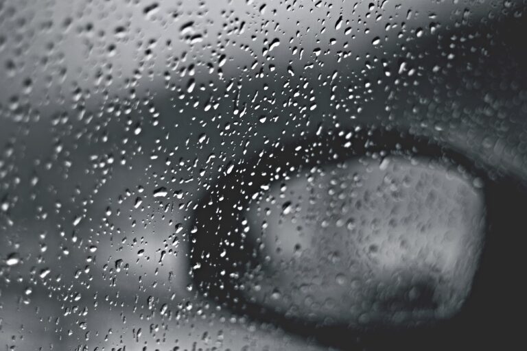 Raindrops on a car window with a blurred side mirror in the background, in black and white.