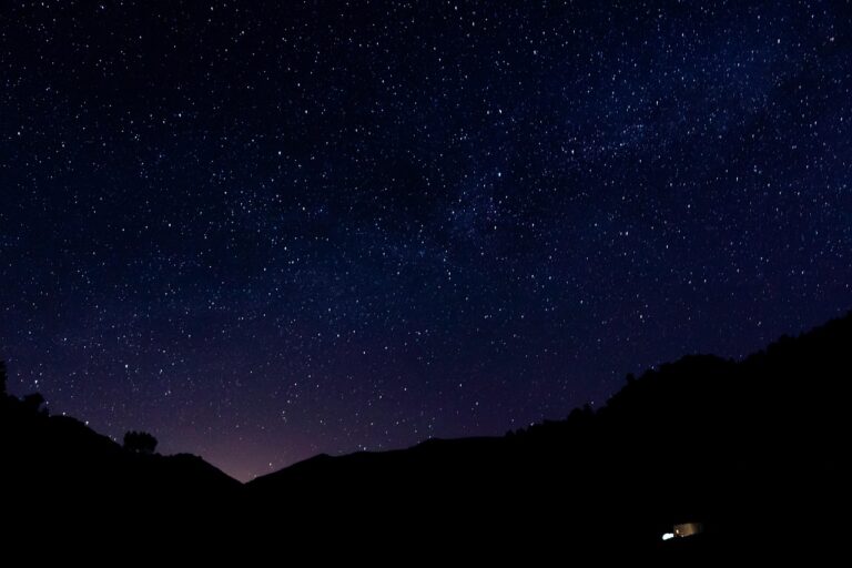 A clear night sky filled with stars above dark silhouettes of hills and a small lit building.