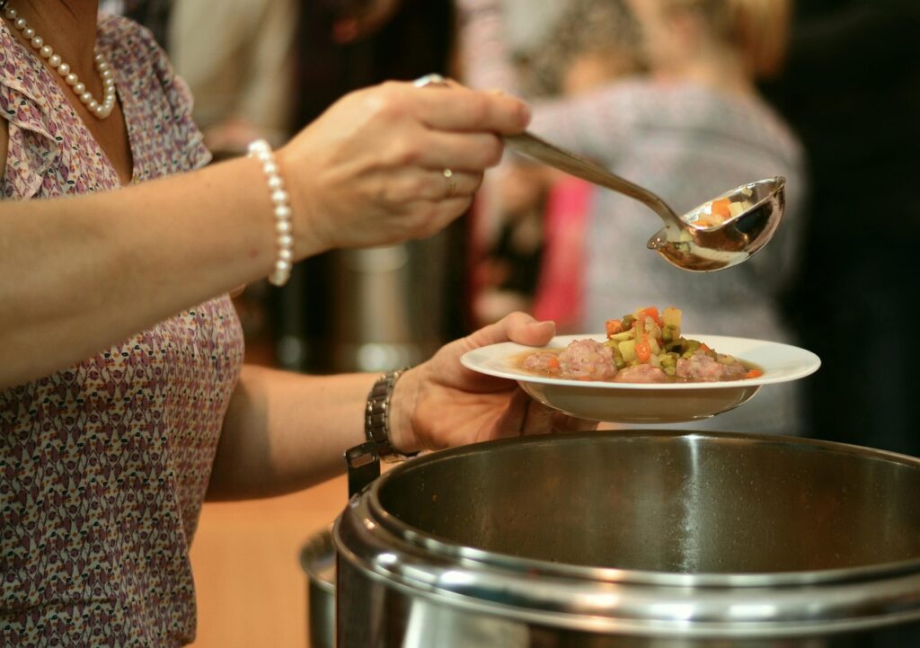 Person serving stew onto a white plate with a ladle at a buffet or soup kitchen.
