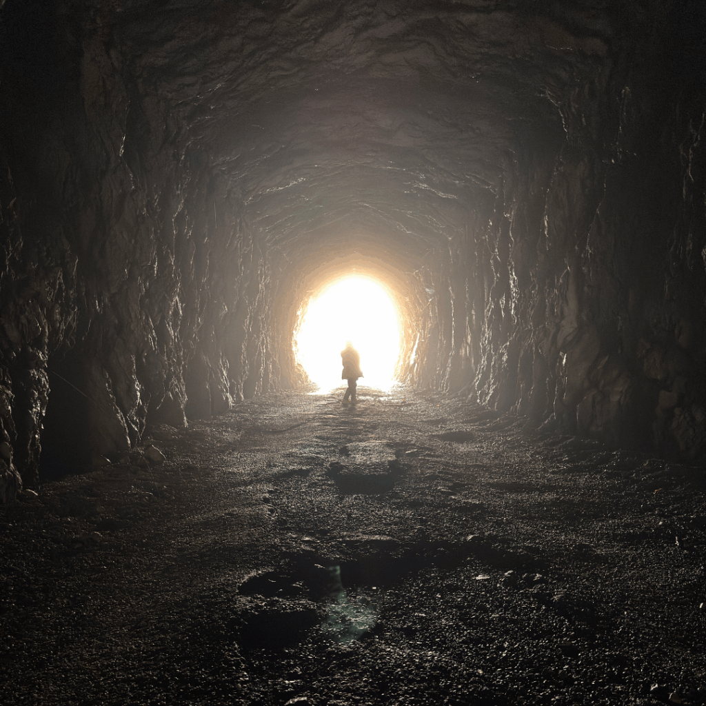 A person stands at the bright entrance of a dark, rocky tunnel.