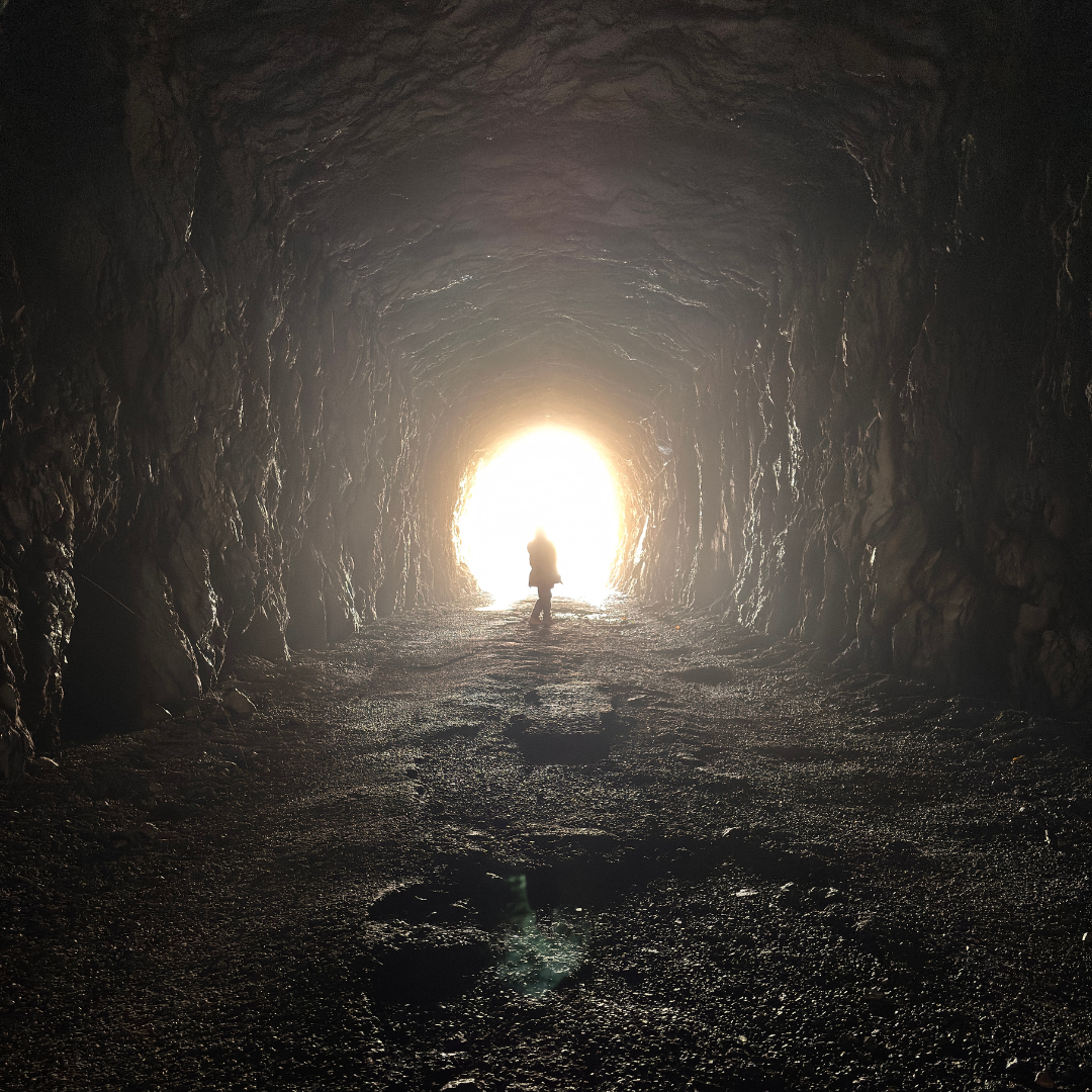 A person stands at the bright entrance of a dark, rocky tunnel.