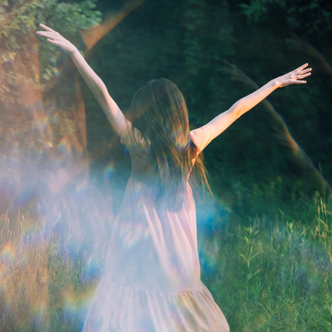 A person in a light dress stands in a sunlit meadow with arms raised, surrounded by soft, dreamy light.