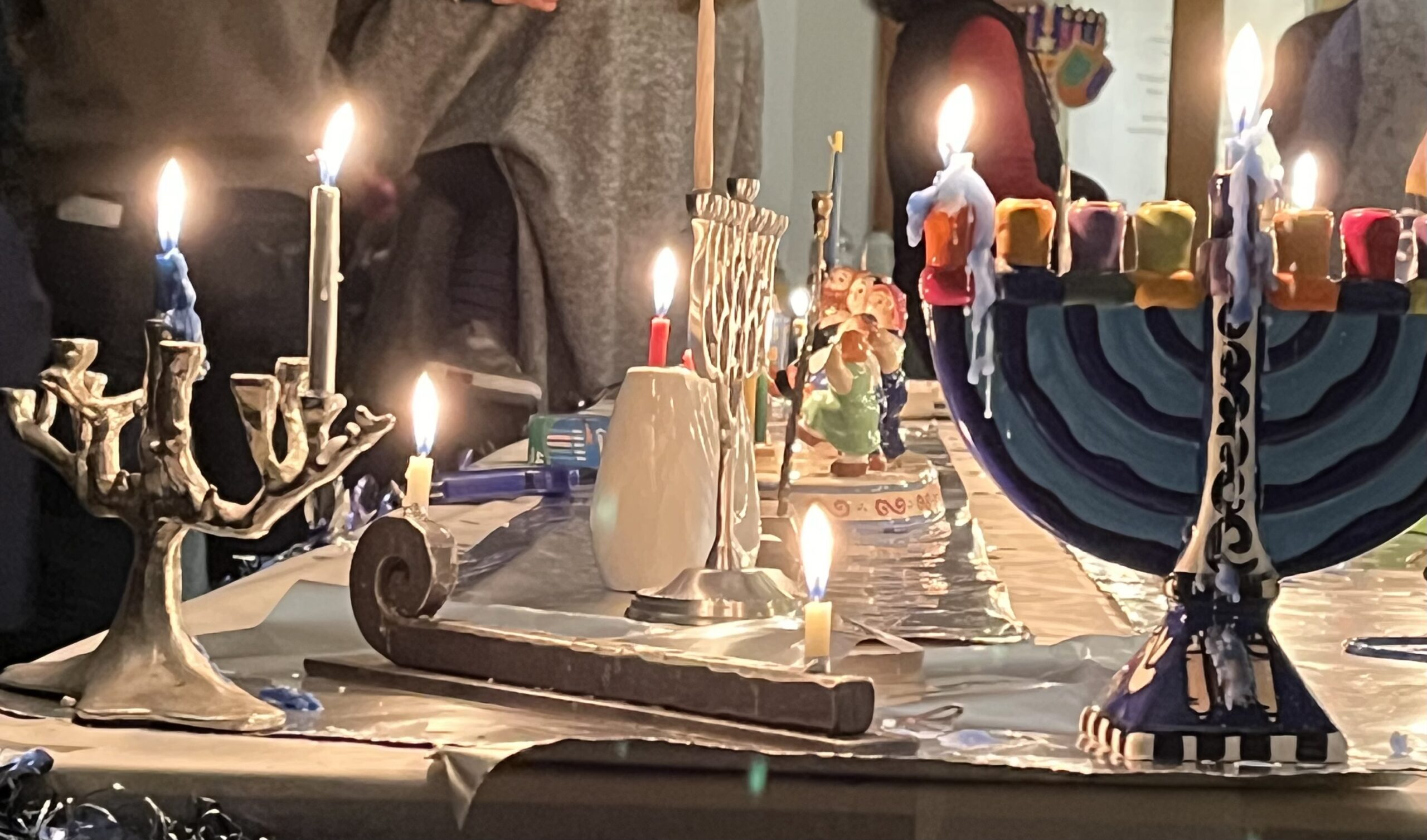 Menorahs and lit candles on a table, celebrating Hanukkah with festive decorations in the background.