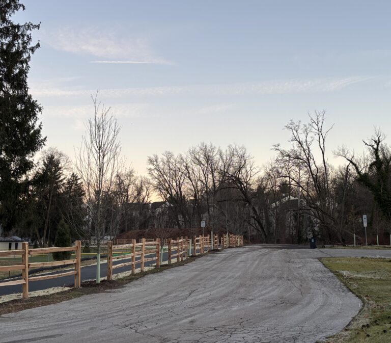 A paved path with a wooden fence curves through a park lined with bare trees at sunset.