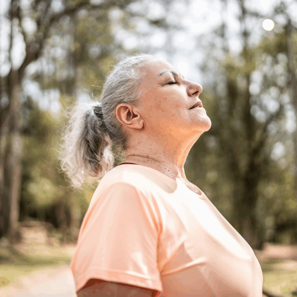 Older woman in a peach shirt stands outdoors with closed eyes, enjoying the fresh air in a sunlit park.