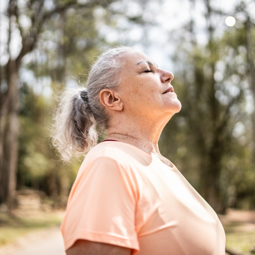 Older woman in a peach shirt stands outdoors with closed eyes, enjoying the fresh air in a sunlit park.