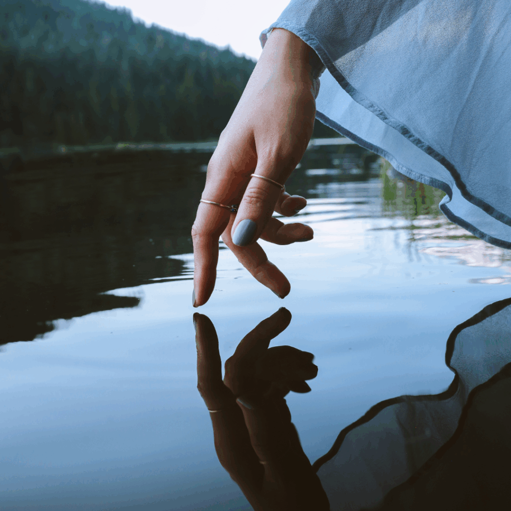 A hand with rings touches calm water, creating ripples, with trees and mountains in the background.