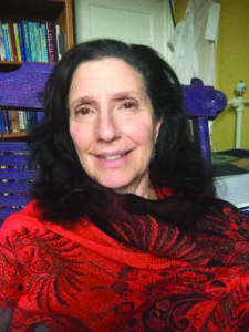 Rabbi Tsurah August is a smiling woman with long dark hair wears a red scarf, sitting indoors in front of a bookshelf.
