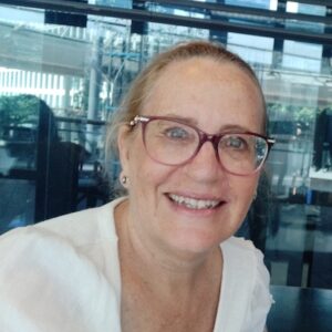 Smiling woman with glasses and a white top sits indoors near a window with blinds.