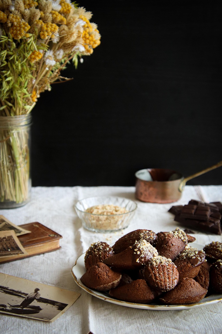 Plate of chocolate madeleines on a table with vintage photos, books, flowers, and dark chocolate pieces.