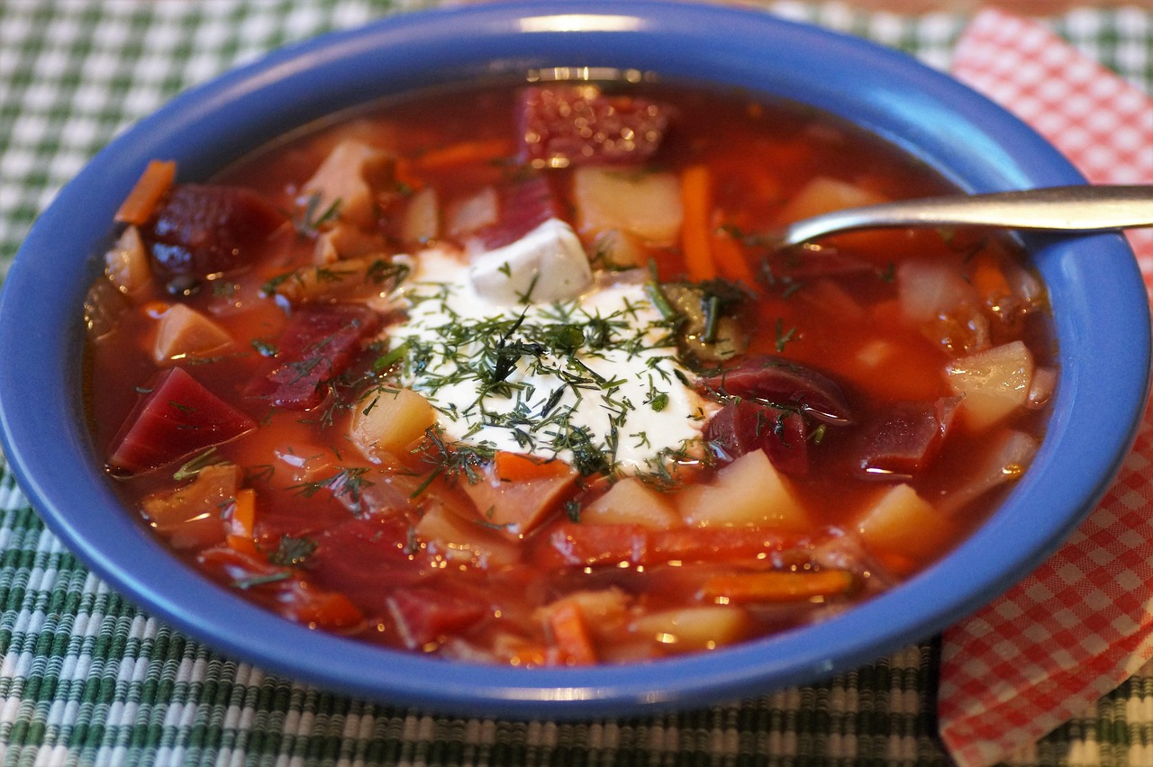 A bowl of borscht soup with sour cream and herbs, served with a spoon on a checkered tablecloth.