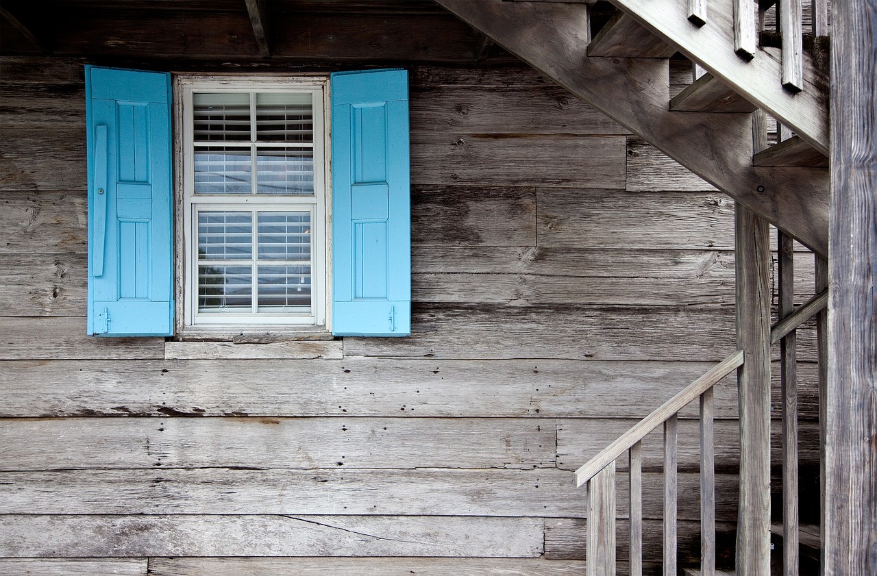 Wooden wall with a window featuring bright blue shutters and a wooden staircase on the right side.