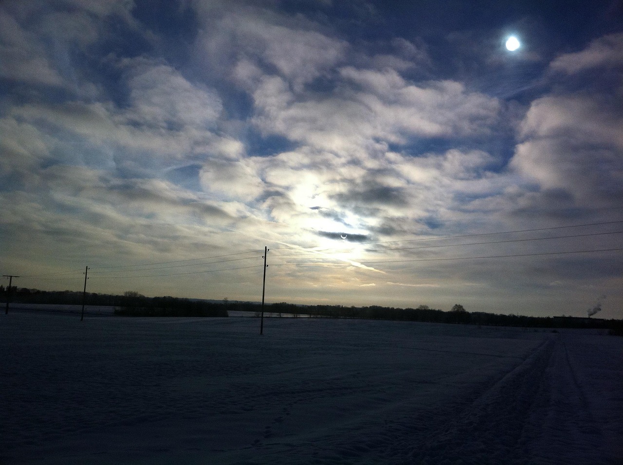 Snowy field under a cloudy sky with the sun and power lines visible in the background.