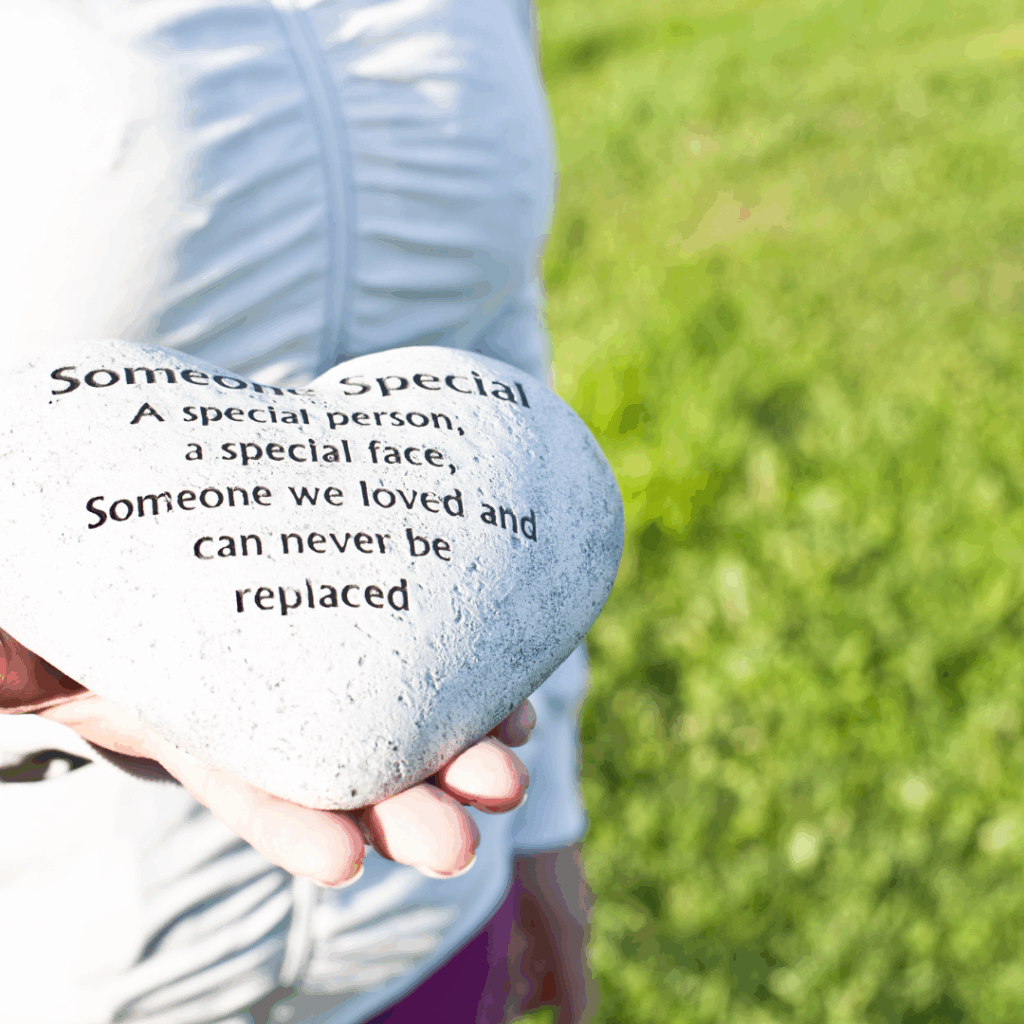 Person holding a heart-shaped stone with a memorial message engraved, standing on green grass.