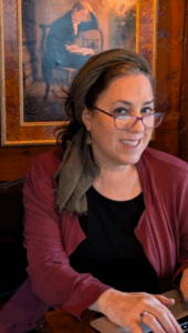 Woman with glasses and headscarf smiling while working on a laptop, with art of a writer in the background.