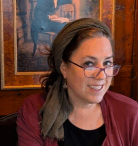 Woman with glasses and headscarf smiling while working on a laptop, with art of a writer in the background.