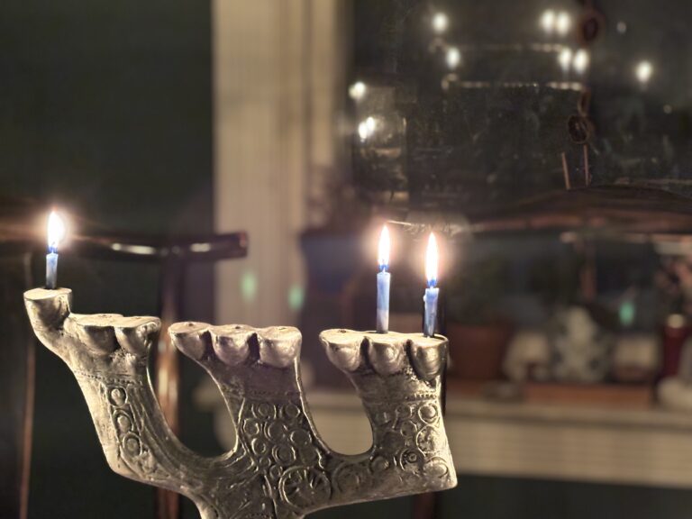 A menorah with two lit candles stands on a table, with a blurred background and mirror reflection.