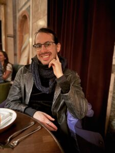 A smiling person with glasses and a scarf sits at a table in a dimly lit restaurant.
