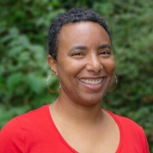 Person with short hair and hoop earrings smiles outdoors, wearing a red top with greenery in the background.