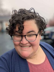 Person with short curly hair, glasses, and a blue sweater smiling outdoors on a cloudy day.