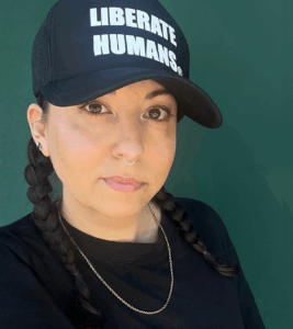 Person in black cap reading LIBERATE HUMANS, black shirt, and braided hair, posing against a green background.