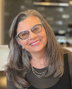 Smiling older woman with glasses and gray hair, wearing hoop earrings and layered necklaces.