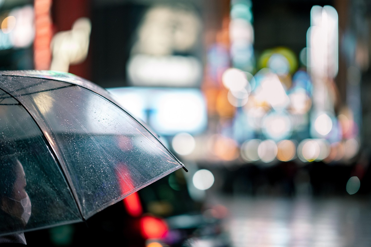 Close-up of a person holding a clear umbrella on a rainy night with city lights blurred in the background.