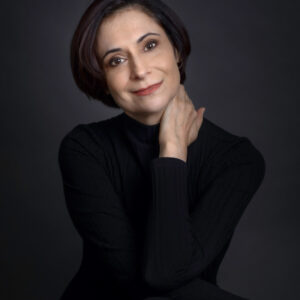 Woman with short dark hair in a black top, smiling softly with one hand touching her neck, on a dark background.