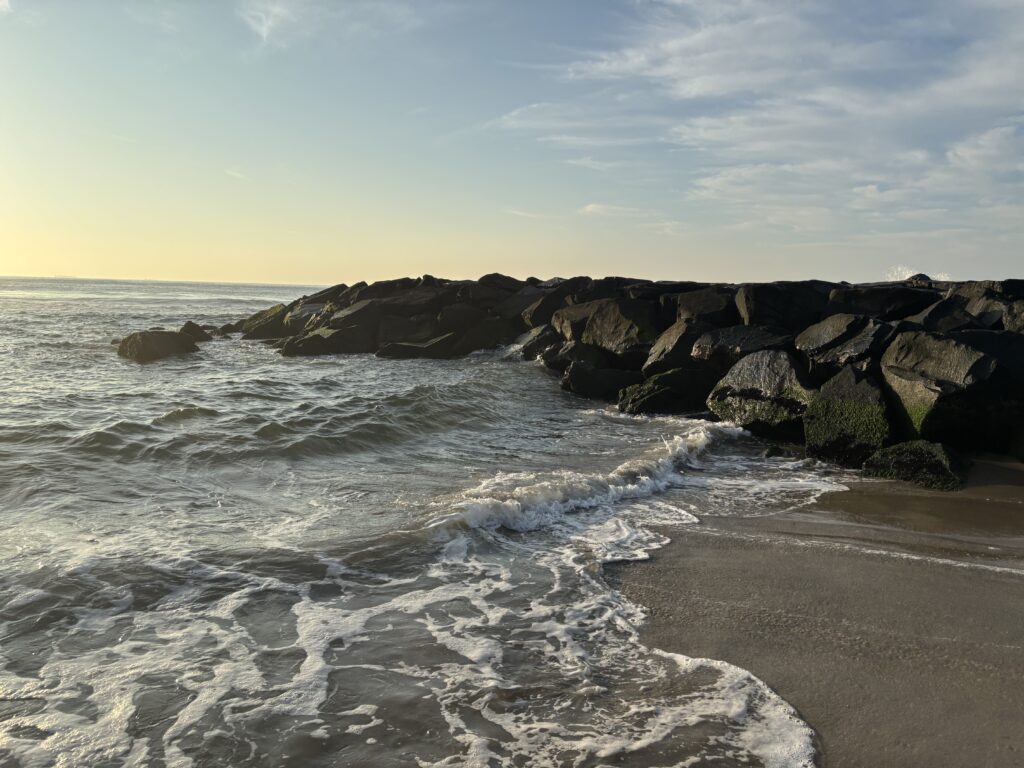 Waves gently washing onto a sandy beach with a rocky jetty under a partly cloudy sky at sunset.