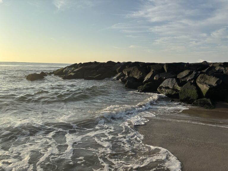 Waves gently washing onto a sandy beach with a rocky jetty under a partly cloudy sky at sunset.