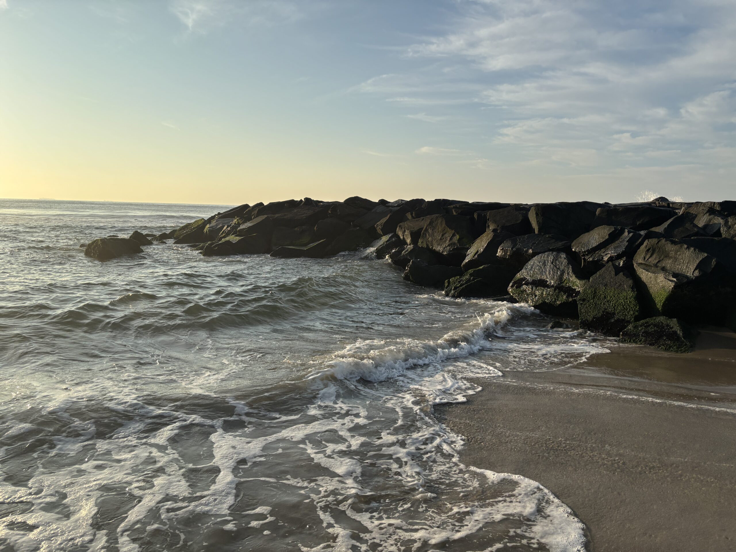 Waves gently washing onto a sandy beach with a rocky jetty under a partly cloudy sky at sunset.