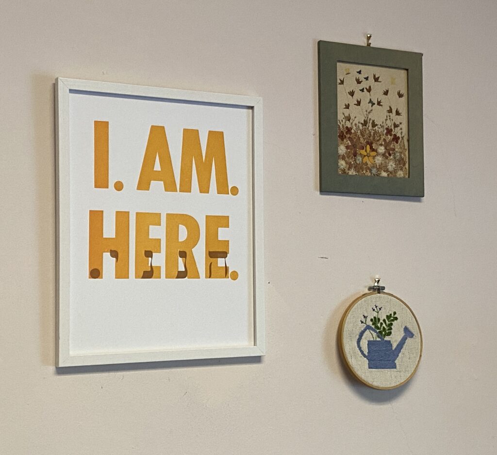 Three framed artworks on a wall, one reads “I AM. HERE.” in bold yellow letters.