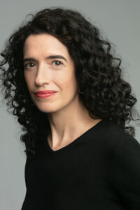 Woman with long, curly dark hair and red lipstick, wearing a black top, against a plain gray background.